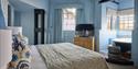 Cosy blue bedroom with a textured bedspread, floral pillows, wood cabinet and TV, plus a sunlit bathroom with freestanding tub beyond.