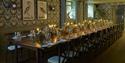Elegant restaurant dining room with a long candlelit table, floral wallpaper, bird artwork, glassware, greenery, and warm festive ambiance.
