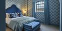 Elegant blue bedroom with botanical wallpaper, scalloped headboard, layered bed, striped bench, teal curtains, and warm bedside lamp.