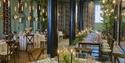 Elegant restaurant dining room with black columns, white-clothed tables, floral centerpieces, lush greenery, and warm pendant lighting.
