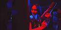 Woman in a dark sci-fi setting holds a futuristic rifle prop, illuminated by vivid red and blue neon; her focused smile creates a dramatic mood.