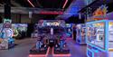 Colorful neon-lit arcade with a motorcycle racing game centered, surrounded by glowing racing cabinets, claw machines, and prize games.