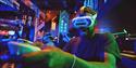 A smiling man wearing a VR headset labeled “HERO” plays an arcade game, illuminated by vivid blue, green, and purple neon lights.