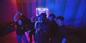 A lively group of five friends performs and dances in a neon-lit recording studio, with a microphone, cassette mural, and vibrant blue-red lights.