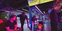 Four friends gather in a vibrant neon-lit arcade, examining colorful bowling balls in a cart beneath glowing “Mega City” signs.