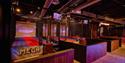 Neon-lit indoor entertainment center with trampoline courts, batting cages, wooden counters, and colorful signs creating an energetic, playful atmosph