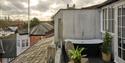 A tranquil rooftop terrace with a black soaking tub, wicker chairs, potted plants, and city rooftops beneath a dramatic cloudy sunset.