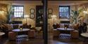 Cosy rustic living room with leather sofas, patterned cushions, plants, lamps, wood floors, and a central black stove pipe; warm, inviting mood.