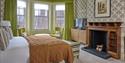 Elegant vintage bedroom with green bay-window curtains, patterned wallpaper, quilted bed, striped chairs, TV cabinet, and wood fireplace.
