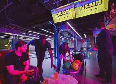 Four friends gather in a vibrant neon-lit arcade, examining colorful bowling balls in a cart beneath glowing “Mega City” signs.